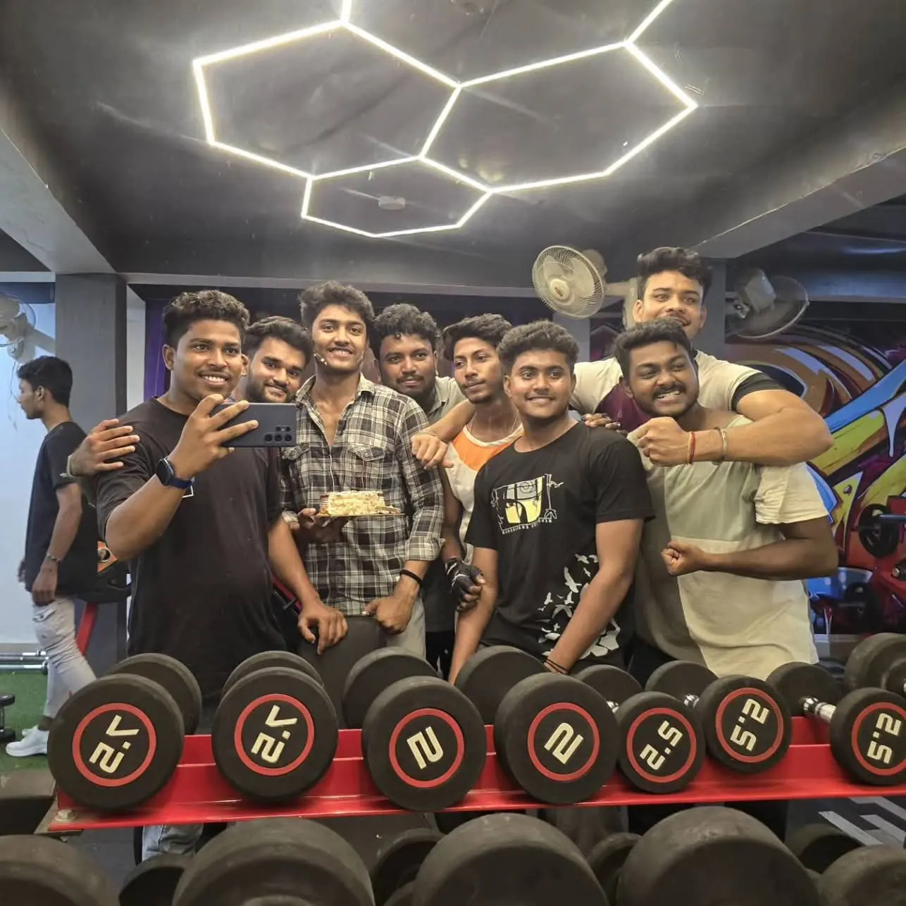 Hexagon LED lights on the ceiling of M.S Fitness above a rack of rubber dumbbells, with members smiling behind it