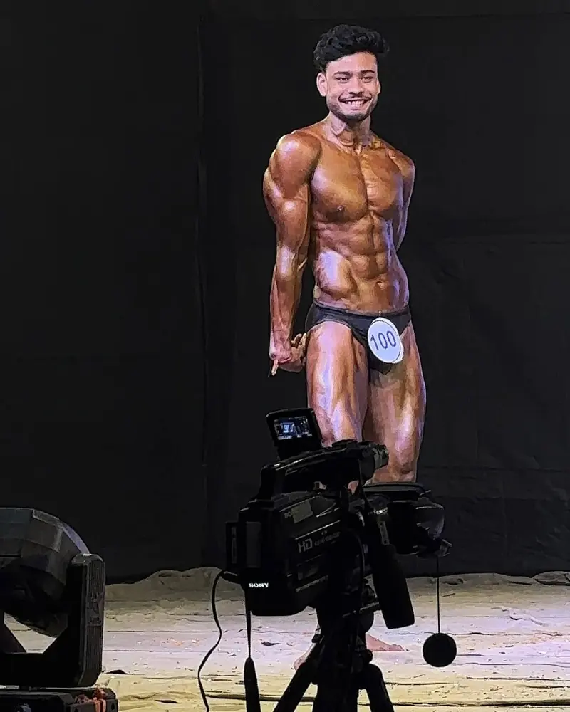 Mayukh smiling on a bodybuilding stage, wearing competitor number 100