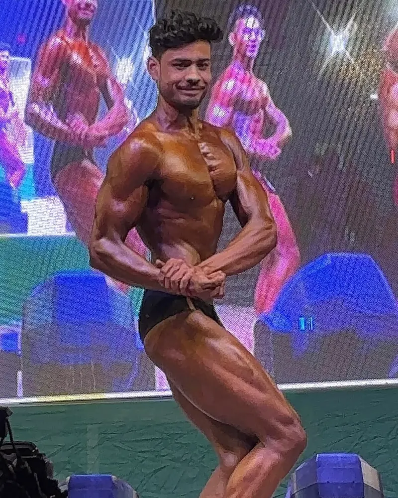 Mayukh hitting a side chest pose on a bodybuilding stage under bright lights